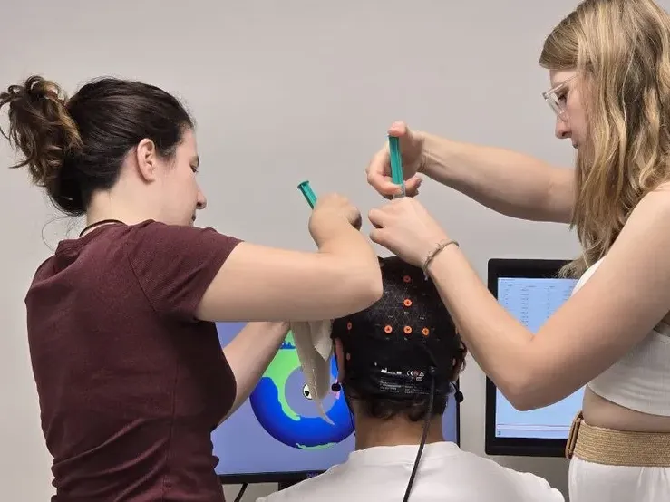 Two student assistants fit an EEG cap on a participant seated in front of a screen.